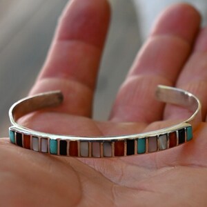 Op de afbeelding: Zilveren manchetarmband met een rij rechthoekige inlays in verschillende kleuren, waaronder turquoise, rood, zwart en wit. De open armband rust op de handpalm van een persoon.