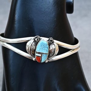Puede incluir: Brazalete de plata con incrustaciones centrales de piedra turquesa, acentuado con patrones geométricos rojos y blancos. El brazalete presenta un diseño de doble banda y detalles decorativos de hojas y rosas.