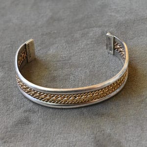 May include: A silver-tone cuff bracelet with a gold-tone braided design. The bracelet has a rectangular clasp and is displayed on a gray surface. The bracelet is a jewelry item.