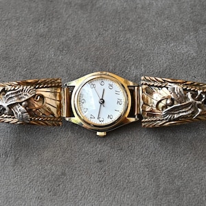 May include: A gold-toned wristwatch with a white face and black numerals. The watch band features detailed, carved designs of birds or eagles. The watch is displayed on a gray surface.