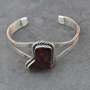 Pode incluir: Uma pulseira de punho prateada com uma pedra retangular castanha em forma de bota de cowboy. A pedra é emoldurada por um design de corda prateada torcida. A pulseira tem um design simples e aberto.