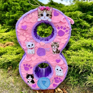 May include: A number eight shaped piñata covered in light pink scalloped tissue paper. It features cutouts of kittens, purple and blue yarn balls, purple paw prints, and small purple hearts. The structure has two circular openings with purple trim.