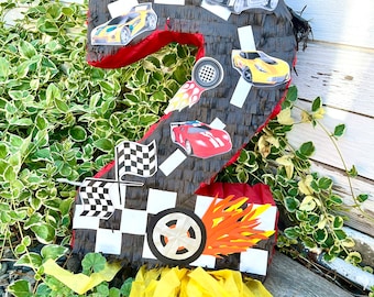 May include: A black and red number two shaped pinata with a chequered race track design and images of racing cars. The pinata has yellow streamers hanging from the bottom.