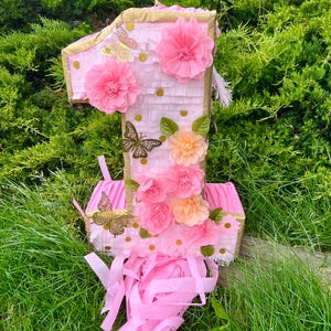 Könnte beinhalten: Eine rosa Piñata in Form der Zahl 1, verziert mit rosa Papierblumen, goldenen Schmetterlingen und goldenen Verzierungen. Die Piñata hat einen rosa Fransenbesatz am unteren Rand, vor grünem Laub und Gras.