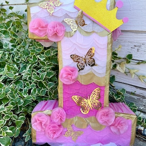 Peut inclure: Une piñata en forme de numéro un, rose et dorée, avec une couronne dorée et des décorations en forme de papillon. La piñata est décorée de papier de soie rose et de paillettes dorées.