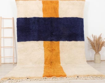 Könnte beinhalten: Ein weißer, blauer und orangefarbener geometrischer Teppich mit Fransen. Der Teppich hat ein großes, klares Kreuzmuster.