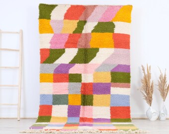 May include: A colorful geometric rug with a white background and squares of pink, green, yellow, orange, blue, and purple. The rug has a fringe border.