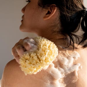 Pode incluir: Uma pessoa está usando uma esponja de luffa natural para lavar o corpo no chuveiro. A esponja é amarela e tem uma superfície texturizada. A pessoa está coberta de espuma de sabão branca.