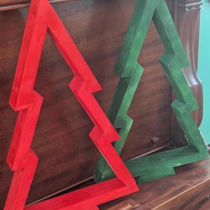 Op de afbeelding: Twee houten kerstboomversieringen. Eén is felrood geschilderd, de andere groen. Elke boom heeft een uniek zigzagontwerp. De bomen worden gepresenteerd op een donker houten oppervlak, wat wijst op een vakantiethema.