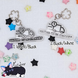 May include: Two clear acrylic keychains with the text "WAITING TO BE ISEKAIED" and a graphic of a truck with a clock. One keychain is white and black, the other is black and white. The keychains are surrounded by colorful star-shaped decorations.