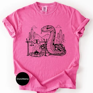 May include: A vibrant pink t-shirt with a black and white illustration of a snake in a desert setting. The snake is positioned near a campfire with a tied-up animal. The text "Crunchberry" is visible on the bottom left of the shirt.