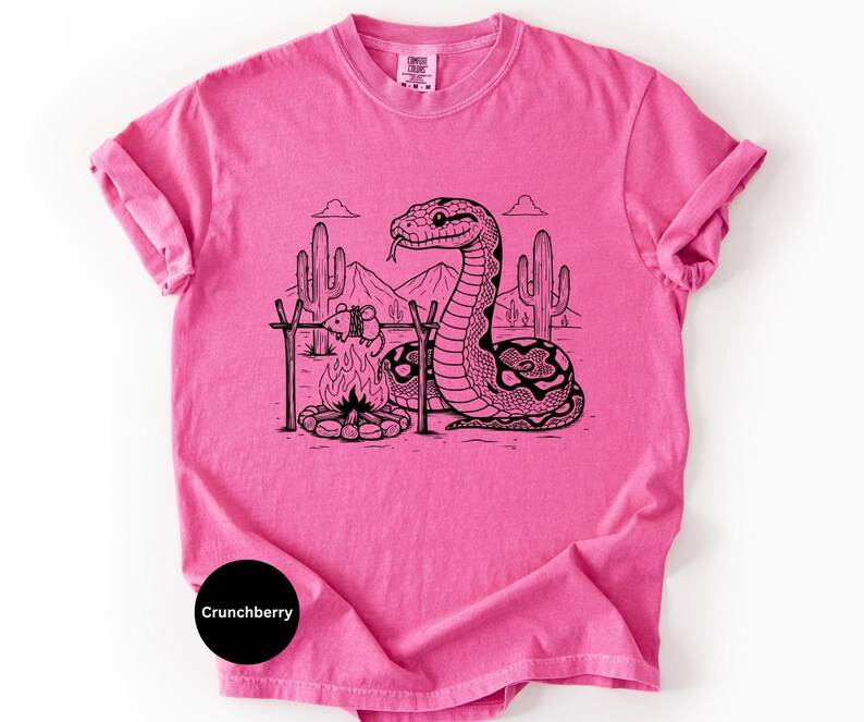 May include: A vibrant pink t-shirt with a black and white illustration of a snake in a desert setting. The snake is positioned near a campfire with a tied-up animal. The text "Crunchberry" is visible on the bottom left of the shirt.