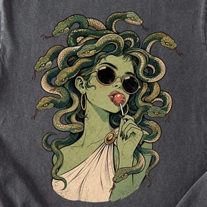 May include: A charcoal gray t-shirt featuring a graphic illustration of a green-skinned figure with snakes for hair, wearing round sunglasses and holding a red lollipop. The figure wears a white draped garment with a gold brooch at the shoulder.