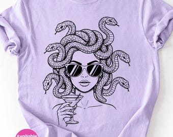 May include: A light purple t-shirt featuring a black and white illustration of Medusa wearing sunglasses and holding a martini glass. The design includes snakes for hair. The text "Available in 5 Colors!" is in a pink circle.