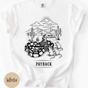 May include: White t-shirt featuring a black and white illustration of a snake with a scarf, mice, and a desert scene. The text "PAYBACK" is printed below the image. The shirt is made of a soft material.