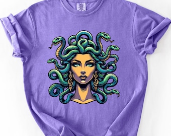 May include: A purple t-shirt featuring a detailed illustration of Medusa with snakes for hair. The artwork showcases a woman's face with teal eyes, gold earrings, and a yellow skin tone. The snakes are green and blue.