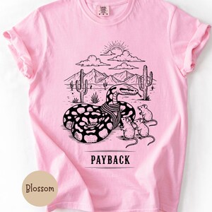 May include: Pale pink t-shirt with a black and white graphic of a snake in a desert scene with cacti and mountains. The text "PAYBACK" is below the graphic. Three mice are near the snake. The shirt has short sleeves.
