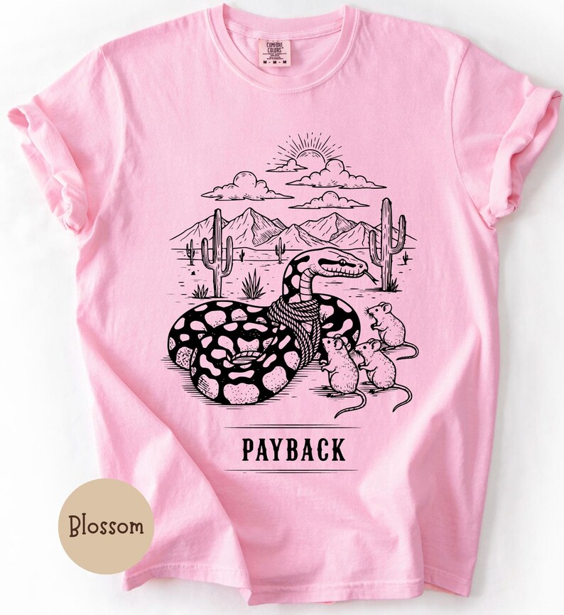 May include: Pale pink t-shirt with a black and white graphic of a snake in a desert scene with cacti and mountains. The text "PAYBACK" is below the graphic. Three mice are near the snake. The shirt has short sleeves.