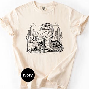 May include: Ivory t-shirt with a black and white desert camping scene featuring a snake, campfire, and cacti. The graphic includes mountains and clouds. The shirt has a crew neck and short sleeves.