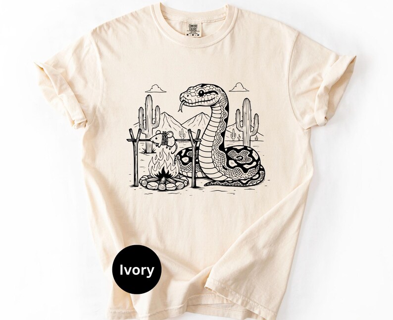 May include: Ivory t-shirt with a black and white desert camping scene featuring a snake, campfire, and cacti. The graphic includes mountains and clouds. The shirt has a crew neck and short sleeves.