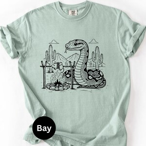 May include: A pale green t-shirt with a black and white graphic of a snake, campfire, and desert scene. The snake is near the fire, with cacti and mountains in the background. The shirt has a crew neck and short sleeves.