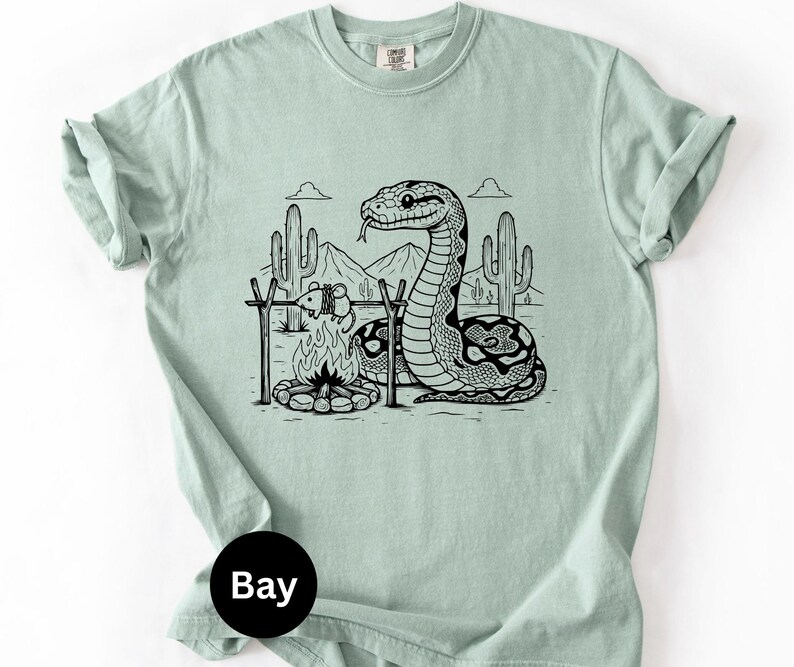 May include: A pale green t-shirt with a black and white graphic of a snake, campfire, and desert scene. The snake is near the fire, with cacti and mountains in the background. The shirt has a crew neck and short sleeves.