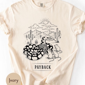 Puede incluir: Camiseta color marfil con un diseño en blanco y negro de un desierto con una serpiente, ratones, cactus, montañas y el sol. La palabra "PAYBACK" está impresa debajo. Marca Comfort Colors.