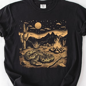 Peut inclure: T-shirt noir avec un motif de paysage désertique. Le graphisme représente un serpent enroulé, un cactus, un feu de camp et des montagnes sous un ciel étoilé et une pleine lune, en tons de marron et d'or.