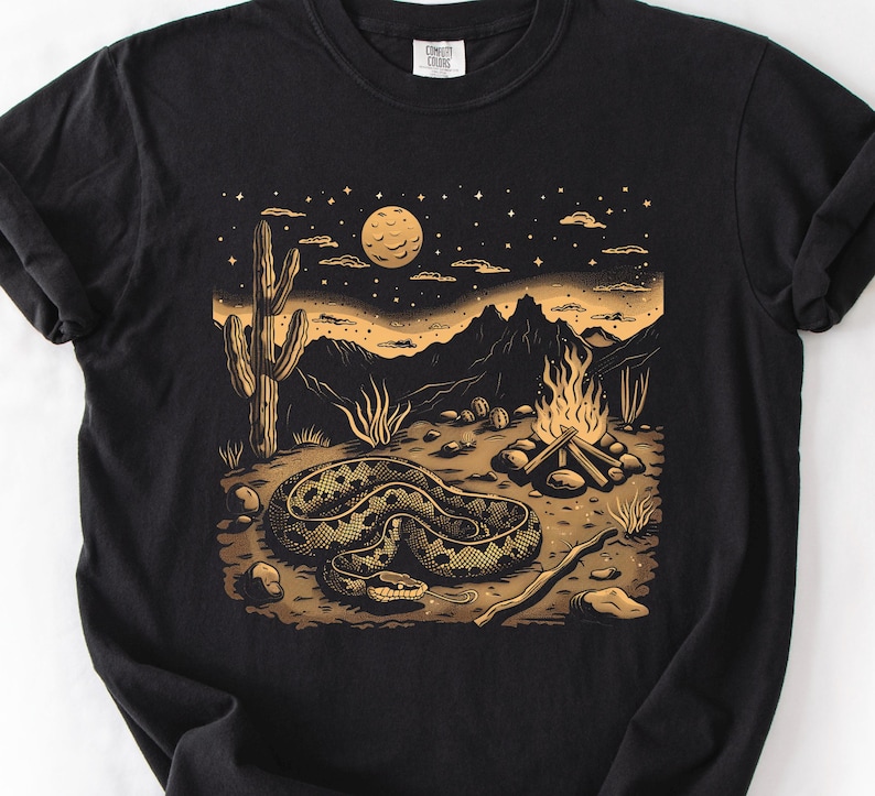 Może przedstawiać: Czarny t-shirt z motywem pustynnego krajobrazu. Grafika przedstawia zwiniętego węża, kaktus, ognisko i g&oacute;ry pod gwiaździstym niebem i pełnią księżyca, w odcieniach brązu i złota.