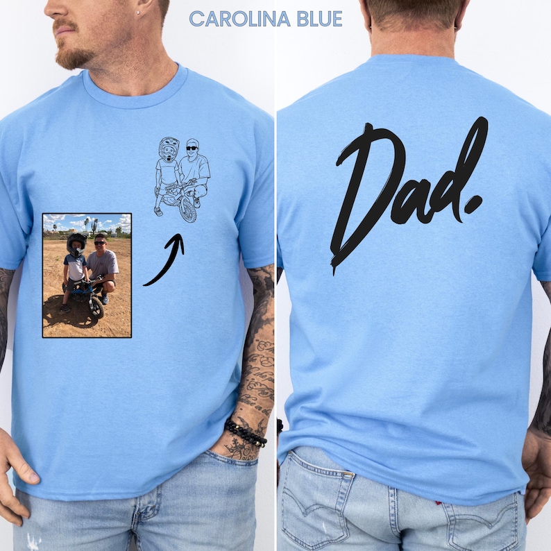 May include: A Carolina blue t-shirt featuring a custom line art illustration on the front chest, derived from a small rectangular photograph of a person and a child on a bike. The back of the shirt displays the word Dad in large black script.
