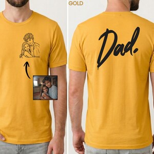May include: A mustard yellow t-shirt featuring a black line art illustration of a person hugging a child on the front left chest. The back of the shirt displays the word Dad in a large black script font. A small photo reference is included.