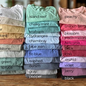 May include: Piles of folded t-shirts in a range of colours, such as white, ivory, butter, mustard, yam, sandstone, bay, light green, blue spruce, and moss. Other colours include island reef, chalky mint, seafoam, hydrangea, chambray, blue jean, flo blue, washed, denim, gray, and pepper.