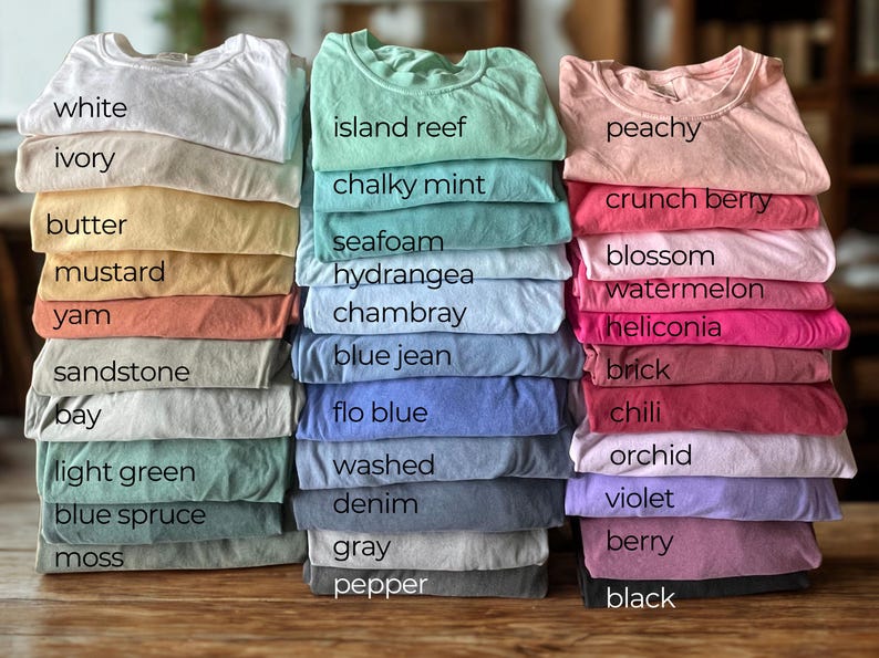 May include: Piles of folded t-shirts in a range of colours, such as white, ivory, butter, mustard, yam, sandstone, bay, light green, blue spruce, and moss. Other colours include island reef, chalky mint, seafoam, hydrangea, chambray, blue jean, flo blue, washed, denim, gray, and pepper.