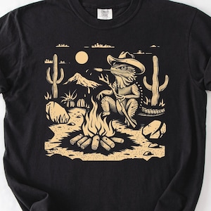 Peut inclure: Un t-shirt noir avec une impression graphique beige clair représentant un lézard portant un chapeau de cowboy, assis près d'un feu de camp dans un désert avec des cactus, des rochers, une montagne et un soleil. Étiquette Comfort Colors au col.