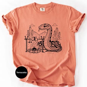 May include: Terracotta-coloured t-shirt with a black and white illustration of a snake camping in a desert scene. The graphic includes a campfire, cacti, and mountains. The shirt has a crew neck and short sleeves.