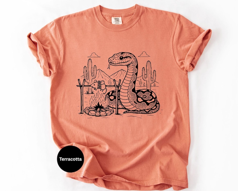 May include: Terracotta-coloured t-shirt with a black and white illustration of a snake camping in a desert scene. The graphic includes a campfire, cacti, and mountains. The shirt has a crew neck and short sleeves.