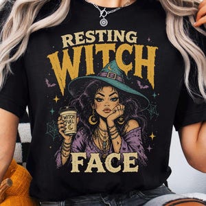 May include: A black t-shirt featuring a graphic of a person wearing a wide-brimmed hat, holding a cup labeled 'WITCH FUEL'. The text 'RESTING' appears above and 'WITCH FACE' below the illustration, which includes stars, bats, and spiderwebs.