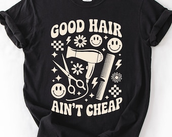 Op de afbeelding: Een zwart T-shirt met korte mouwen en een crèmekleurige grafische print. Het ontwerp bevat een haardroger, een schaar, een kam, smileys, bloemen, bliksemschichten en dambordpatronen met de tekst GOOD HAIR AIN'T CHEAP.