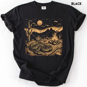 Może przedstawiać: Czarny t-shirt z motywem pustynnego krajobrazu. Ilustracja przedstawia węża, ognisko, kaktus i g&oacute;ry pod rozgwieżdżonym niebem. Grafika jest w odcieniach beżu i brązu, z napisem "BLACK" u g&oacute;ry.