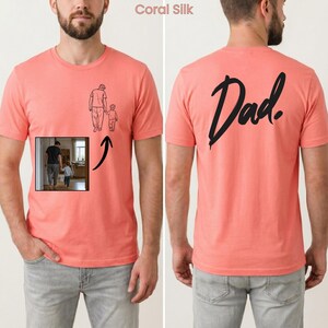 May include: A coral silk t-shirt featuring a black line drawing of an adult holding a child's hand on the front left chest. The back of the shirt displays the word Dad in a large, black, handwritten-style script font.