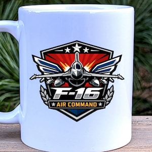 May include: White ceramic mug featuring a detailed graphic of an F-16 fighter jet. The design includes the text "F-16 AIR COMMAND" within a shield-shaped emblem with stars and wings. The mug has a standard handle.