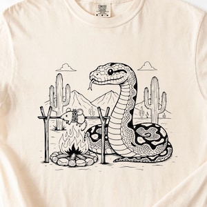 Puede incluir: Camiseta color crema con una ilustración en blanco y negro de una serpiente, una fogata y un paisaje desértico. La serpiente está cerca del fuego, con un ratón asándose. La camiseta tiene una etiqueta Comfort Colors.