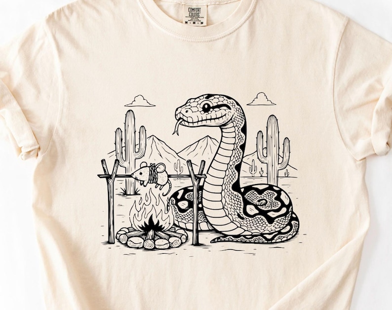 May include: Cream-coloured t-shirt with a black and white illustration of a snake, campfire, and desert landscape. The snake is near the fire, with a mouse roasting. The shirt has a Comfort Colors tag.