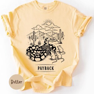 May include: Pale yellow t-shirt featuring a black and white graphic of a snake in a desert scene, with the word "PAYBACK" underneath. The snake has a rope around its neck, and is surrounded by cacti, mountains, and three mice.