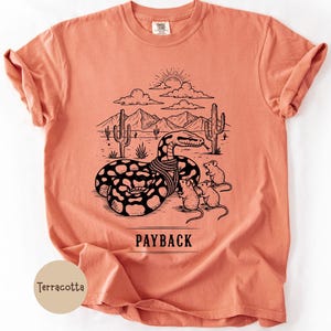 May include: Terracotta t-shirt with a black and white graphic of a snake in front of a desert scene with cacti, mountains, and mice. The word "PAYBACK" is printed below the graphic.