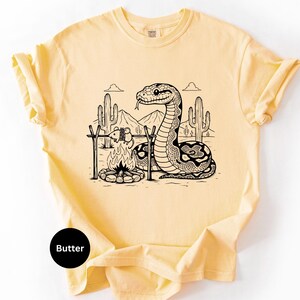 May include: Pale yellow t-shirt with a black and white desert scene print. The graphic depicts a snake, campfire, and mouse, with cacti and mountains. The word "Butter" is printed on a black circle. The t-shirt is a light, neutral colour.