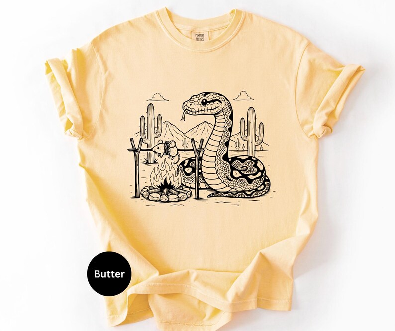 May include: Pale yellow t-shirt with a black and white desert scene print. The graphic depicts a snake, campfire, and mouse, with cacti and mountains. The word "Butter" is printed on a black circle. The t-shirt is a light, neutral colour.