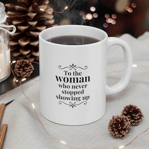 Könnte beinhalten: Weiße Keramiktasse mit schwarzer Aufschrift: „To the Woman who never stopped showing up“. Die Tasse hat einen gebogenen Henkel und ist mit einer dunklen Flüssigkeit gefüllt. Dekorative Elemente sind Tannenzapfen und Lichterketten.