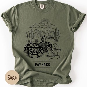May include: Sage green t-shirt featuring a black graphic of a snake in a desert scene with cacti, mountains, and clouds. Three mice are near the snake. The word "PAYBACK" is printed below the graphic.