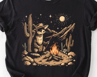 May include: A black t-shirt featuring a graphic illustration of an otter wearing a cowboy hat, sitting by a campfire in a desert landscape with cacti, mountains, and a starry night sky. The Comfort Colors brand label is visible at the neckline.